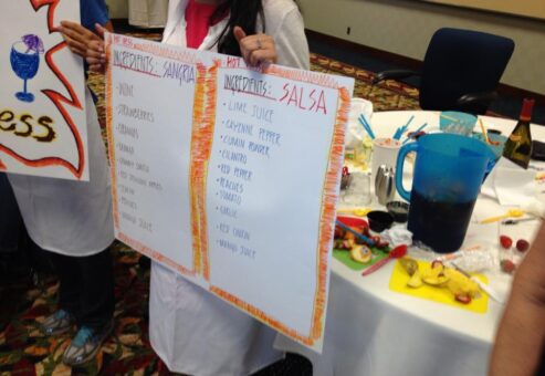A team member holds up ingredient lists for Sangria and Salsa at the Salsa Showdown team building event, with colorful ingredients and materials on the table.
