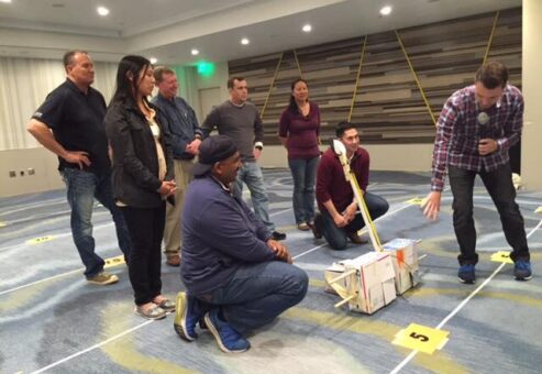 A group of participants indoors discussing their catapult design as part of the Catapult to Success team building activity, focusing on teamwork, strategy, and collaborative problem-solving in a competitive setting.