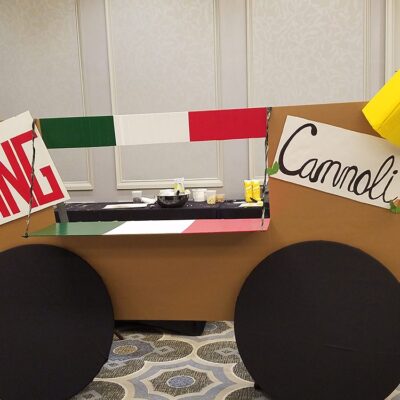 An imaginative 'food truck' display titled 'King Cannoli' is set up as part of a team building event. The mock food truck is constructed with a cardboard facade, and the colors of the Italian flag decorating the upper edge. This creative setup reflects the team's effort to embody an Italian pastry theme for the 'Food Truck Challenge'. thumbnail