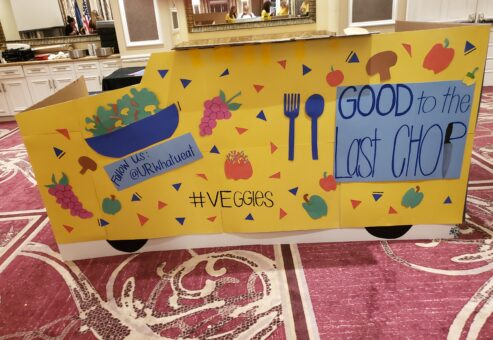 A colorful, handmade food truck façade created for the 'Food Truck Challenge' team building event. The bright yellow design features cut-out images of fruits and vegetables, along with slogans like 'Good to the Last Chop'. The creative display emphasizes a healthy, veggie-focused theme, highlighting the event's blend of culinary skills and artistic expression.