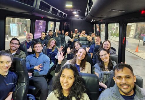 A group of smiling participants sits inside a bus, waving at the camera during a Do Good Bus team building event. The participants are dressed casually, ready to engage in a community service activity.