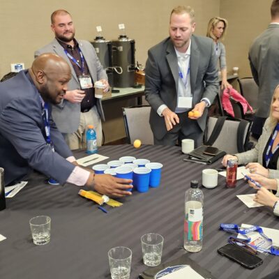 Group of professionals participating in a corporate team building event. One man arranges blue cups on the table while others watch and engage in discussion. thumbnail