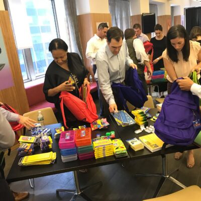 Corporate team members packing backpacks with school supplies during a csr team building event. The table is filled with notebooks, pencils, and other educational materials as participants work together to prepare donations, emphasizing teamwork and giving back to the community. thumbnail