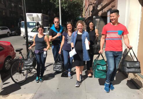 A group of six people walking down a city sidewalk, smiling and carrying insulated bags. They are engaged in a team building activity, possibly delivering meals or supplies.