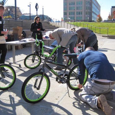 Group of participants in an outdoor Charity Bike Build team building event. Some members are kneeling to work on the bikes, while others stand nearby, discussing instructions. The event is taking place in an open urban space. thumbnail