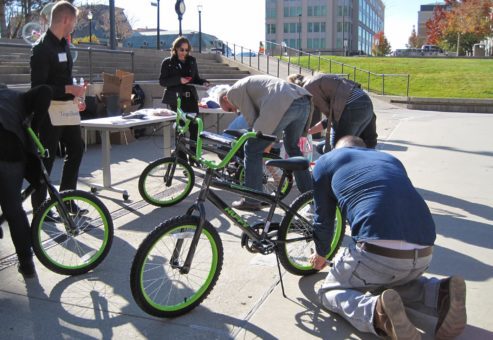 Group of participants in an outdoor Charity Bike Build team building event. Some members are kneeling to work on the bikes, while others stand nearby, discussing instructions. The event is taking place in an open urban space.
