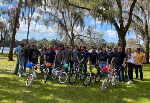 Large group of participants posing outdoors with completed bikes from a Charity Bike Build event. Each assembled bike is decorated with colorful balloons. Participants are smiling in a celebratory mood after a successful team building activity.