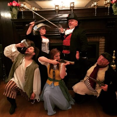 A group of actors in pirate-themed costumes pose dramatically for a Murder Mystery Dinner event. The characters include a pirate captain in a black hat and red vest, a woman dressed as a swashbuckler holding a sword, another woman in a long blue and yellow dress aiming a musket, and two additional pirates—one with a mug and the other with a flintlock pistol. They stand in front of an elegant fireplace, creating a lively and adventurous scene. thumbnail