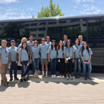 A group of fourteen people standing in front of a large black bus, all wearing matching light blue t-shirts, participating in a team building event. The individuals are smiling and standing close together, indicating camaraderie and a shared purpose. thumbnail