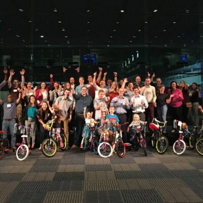 Group of participants smiling and cheering indoors during a Charity Bike Build event. In the front, children excitedly stand with their newly built bikes, surrounded by colorful balloons. The event celebrates teamwork and community engagement as the group successfully completes the charity project. thumbnail