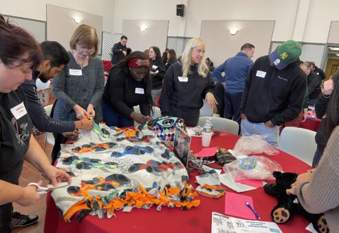 Team members actively collaborate to create cozy blankets during a Hope for the Holidays team building event, contributing to gift baskets that will bring warmth and joy to seniors this holiday season.
