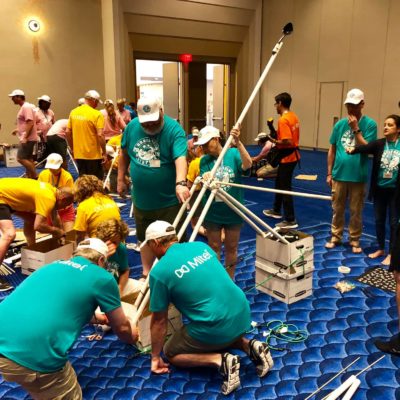 Multiple teams in brightly colored shirts collaborating on building catapults indoors during the Catapult to Success team building activity, showcasing teamwork, creativity, and hands-on construction skills in a lively group setting. thumbnail