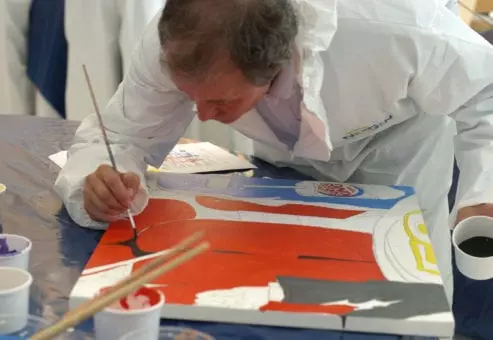 A participant wearing a white protective suit carefully paints a section of a mural during The Big Picture team building event. The individual focuses on adding details to a vibrant red, blue, and white canvas, contributing to the collaborative masterpiece.