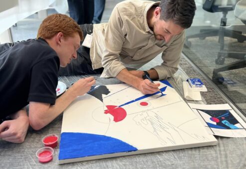 Two participants are lying on the floor, working together to paint a section of the canvas during The Big Picture team building event. They focus intently on adding details and colors, collaborating on their contribution to the larger mural.