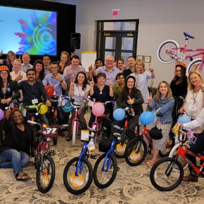 Large group of smiling participants gathered around a collection of brightly colored children's bicycles during a Charity Bike Build event. Many participants are holding up the bikes they built, decorated with balloons and streamers, showcasing the teamwork and joy of the event. thumbnail