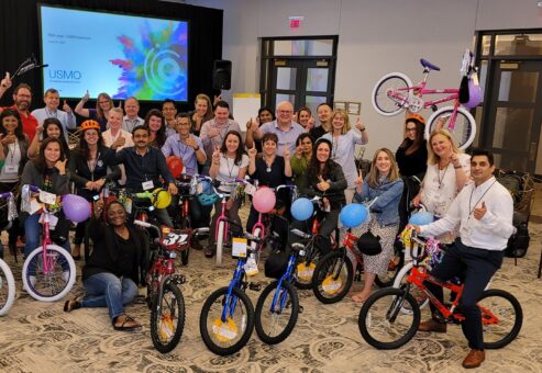 Large group of smiling participants gathered around a collection of brightly colored children's bicycles during a Charity Bike Build event. Many participants are holding up the bikes they built, decorated with balloons and streamers, showcasing the teamwork and joy of the event.