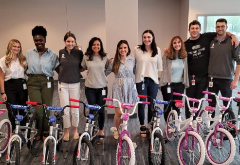 A group of smiling participants standing behind a row of assembled children's bicycles during a Charity Bike Build event. The bikes are pink and blue with training wheels, and the group appears happy with their completed team project.