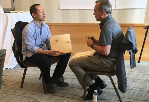 Two participants sit facing each other, discussing their wooden boards during a transformational team building experience centered on breaking through barriers.
