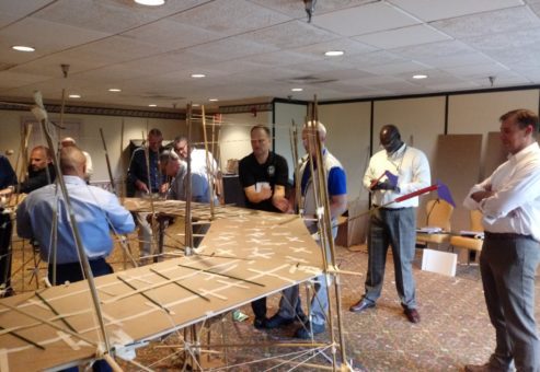 Participants in the Bridging the Divide team building activity gather around a large, collaboratively built cardboard bridge, assessing its structure and stability.