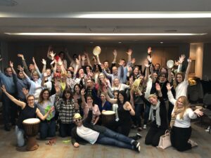 A large group of participants celebrating together after a successful BeatsWork team building session, holding percussion instruments and posing with enthusiasm indoors.