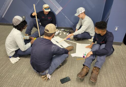 A group of participants sitting on the floor indoors, discussing and planning their catapult design during the Catapult to Success team building activity, emphasizing teamwork, collaboration, and strategic thinking.