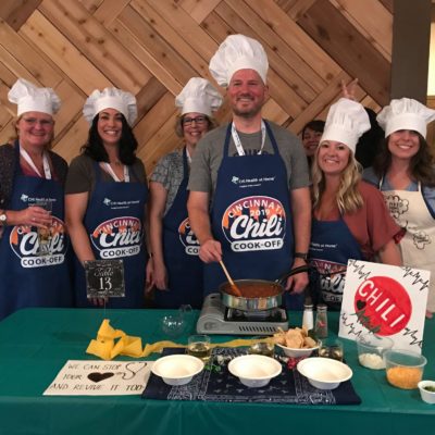 A group of seven people stand behind a table, smiling and wearing chef hats and aprons for a Chili Cook-Off event. They are preparing a chili dish with a pot on a portable stove in front of them. Various ingredients and garnishes are laid out on the table. A sign reads 'We can stop your heart and revive it too!' indicating a playful theme. The group is enjoying a team building culinary event. thumbnail
