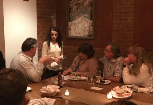 A group of four people sitting around a dimly lit dinner table engaged in conversation, with a female actor standing and looking at one of the seated individuals. The setting suggests an intimate dining experience, part of a Murder Mystery Dinner event, as the group interacts with the actor in the unfolding mystery.