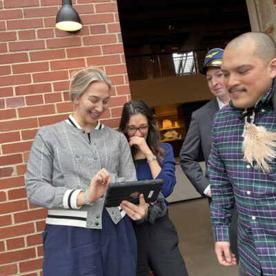 A group of colleagues smiling and interacting with a tablet during an outdoor team building exercise, demonstrating collaboration and teamwork while enjoying the making a movie activity. thumbnail