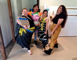 Four participants in a team building activity are standing together, proudly showing off their creative outfits made from recyclable materials. The person on the left wears a black trash bag with yellow 'Caution' tape and an egg carton as part of the design. The second person is dressed in a colorful, patchwork-like outfit with orange spikes made from recycled materials on the shoulders. The third person has a similar 'Caution' tape dress with a black and white design. The fourth person stands confidently in tan pants and black platform shoes, smiling at the camera.