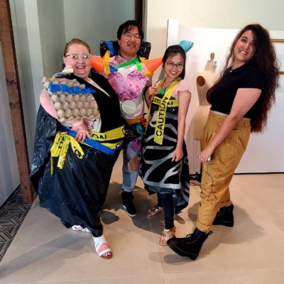 Four participants in a team building activity are standing together, proudly showing off their creative outfits made from recyclable materials. The person on the left wears a black trash bag with yellow 'Caution' tape and an egg carton as part of the design. The second person is dressed in a colorful, patchwork-like outfit with orange spikes made from recycled materials on the shoulders. The third person has a similar 'Caution' tape dress with a black and white design. The fourth person stands confidently in tan pants and black platform shoes, smiling at the camera. thumbnail
