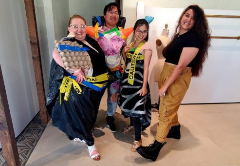 Four participants in a team building activity are standing together, proudly showing off their creative outfits made from recyclable materials. The person on the left wears a black trash bag with yellow 'Caution' tape and an egg carton as part of the design. The second person is dressed in a colorful, patchwork-like outfit with orange spikes made from recycled materials on the shoulders. The third person has a similar 'Caution' tape dress with a black and white design. The fourth person stands confidently in tan pants and black platform shoes, smiling at the camera.