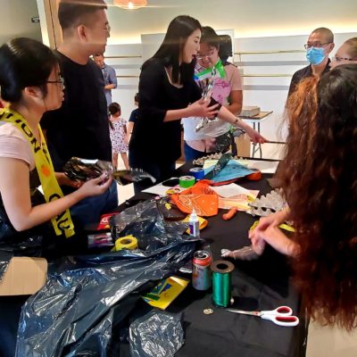 A group of participants engages in a Corporate Project Runway team building activity centered around creating fashion items. Several people are gathered around a table filled with various crafting materials such as scissors, ribbons, and recyclable items. Some participants are cutting and arranging materials while others are brainstorming or collaborating. One person wears an outfit made of trash bags and caution tape, reflecting the creative and playful nature of the event. thumbnail