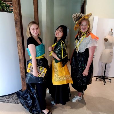 Three women standing together, smiling and posing for a photo during a team building activity. They are dressed in creative outfits made from various unconventional materials like trash bags, plastic, caution tape, and fabric. One of the women wears a large decorative hat made from a mix of recyclable materials. thumbnail