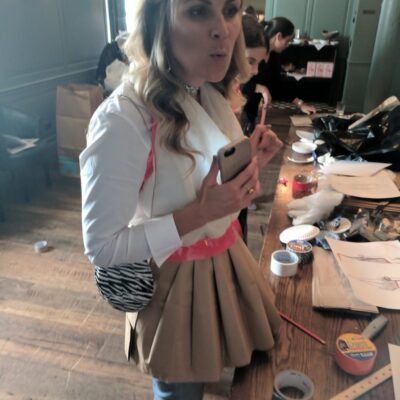A woman standing near a table full of crafting materials, wearing a creative outfit made of paper or fabric for a team building event. Another participant is seen working in the background, focused on the materials laid out. thumbnail