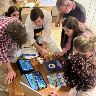 Participants in the 'Corporate CSI' team building challenge gather around a table, focused on solving clues from their detective briefcases and using digital tools to navigate a virtual crime scene. thumbnail