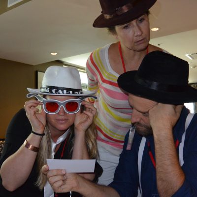 A team participating in the Escape the Mob event focuses on solving clues, while wearing fun mobster-themed hats and sunglasses to fit the robbery mystery challenge. thumbnail