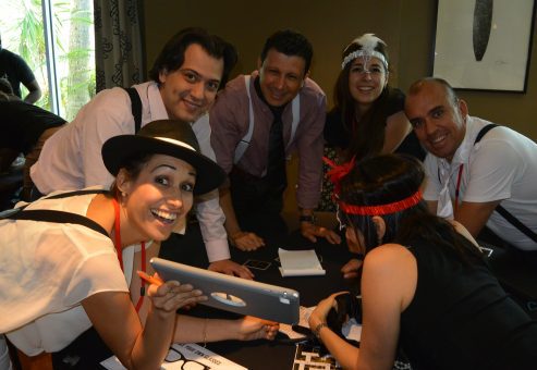 A group of participants in the Escape the Mob team building event works together, using a tablet to solve clues and track down the stolen diamonds while embracing their mobster-themed costumes.