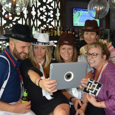 Group of five people in hats smiling and looking at a tablet during a team building activity at a bar, participating in the 'Escape the Mob' program. They are engaged in solving a scavenger hunt and murder mystery challenge, creating a fun and interactive atmosphere. thumbnail