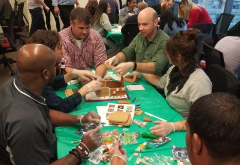 A team of six participants gathered around a table covered with a green cloth, actively building a gingerbread house during a team building competition. The group is engaged in the process, wearing gloves, and using various candy and gingerbread materials to assemble their house. Other teams can be seen in the background participating in the same activity in a lively and collaborative setting.