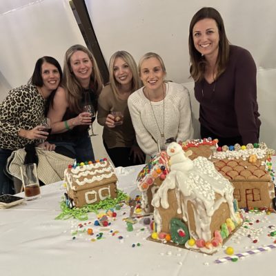 Five smiling participants gather around a table displaying their decorated gingerbread houses during a team building event. The gingerbread houses are intricately adorned with icing, candy, and festive decorations, including a snowman figure on one of the houses. thumbnail