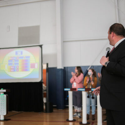 A game show host holding a microphone and looking at a large screen displaying the game board, while participants cheer and clap during the Corporate Feud team building activity in a gymnasium setting. thumbnail