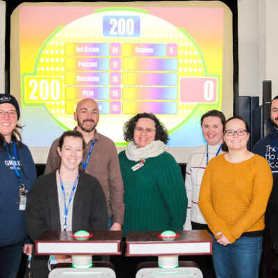 A group of eight participants smiling and standing together in front of a game show setup with a brightly colored game board in the background during the Corporate Feud game. thumbnail