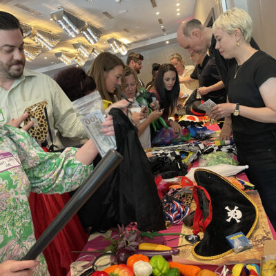 A group of people enthusiastically selecting costumes and props from a table filled with various items like pirate hats, colorful fabrics, and accessories during a team building activity focused on creativity and fun role-playing. thumbnail