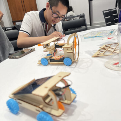 A participant focused on assembling a miniature car from a STEM kit during the STEM Speedway team building activity, with completed wooden model cars on the table, highlighting precision, creativity, and hands-on learning. thumbnail