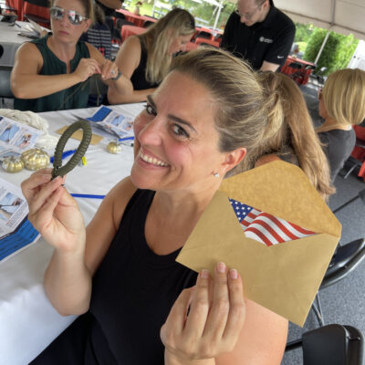Smiling woman holding an envelope with an American flag and a small parcord bracelet during a corporate team building event, with other participants working in the background. thumbnail
