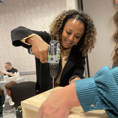 A woman smiles as she uses a power drill to assemble a wooden box, with another person holding the piece steady during the process. thumbnail