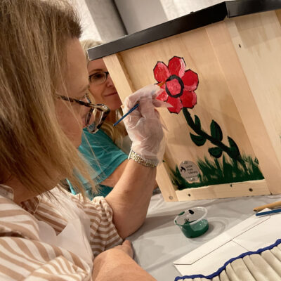 Participant painting a red flower on a wooden Little Free Library as part of the Little Team Library team building event, where teams assemble and decorate book exchange boxes for the community. thumbnail