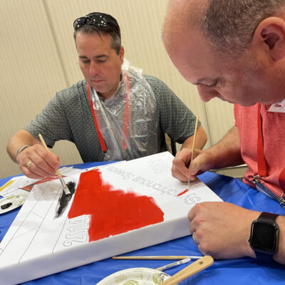 Two men carefully paint a section of a mural during The Big Picture team building event. They are focused on adding red and black colors to their canvas, with a color wheel and brushes nearby. thumbnail