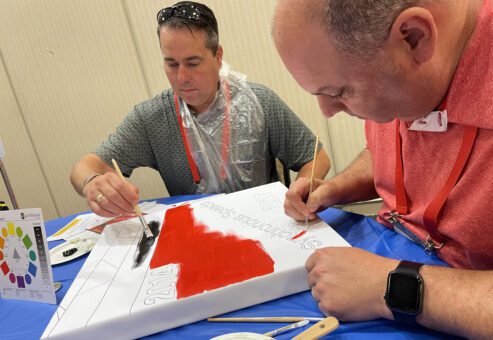 Two men carefully paint a section of a mural during The Big Picture team building event. They are focused on adding red and black colors to their canvas, with a color wheel and brushes nearby.
