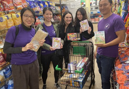 A group of five volunteers stands together in a grocery store aisle, smiling and holding up various food items. They have a shopping cart filled with packaged goods for a donation or charity event. They are participating in a Do Good Bus team building event where they are collecting items to support a community cause. The atmosphere appears cheerful and collaborative.